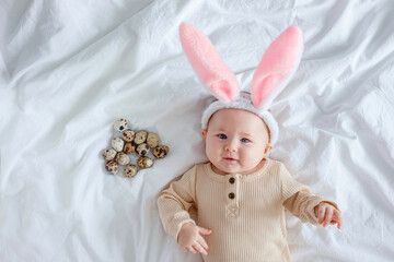A cute funny baby with bunny ears costume dressed up for Easter laying on white bed linen sheet with quail eggs. An infant looks at the camera. Easter costume for children.