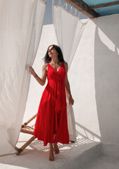 vacation mood. brunette woman in red summer dress stands near white cloth on the white wall background at sunny day. greek villa. travel concept, free space