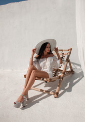 vacation mood. brunette woman with long legs in white summer dress and white hat sits on a beach...