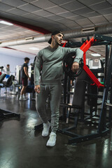 Obraz premium Chico joven musculoso con chándal gris y gorra negra posando en un gimnasio rodeado de maquinas de musculación 