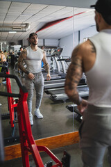 Chico tatuado musculado mirándose y posando delante de un espejo en el gimnasio