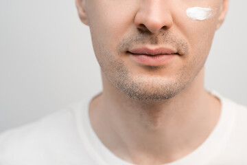 A handsome man with a stubble in a white t-shirt, a sample of a face cream on his cheek.