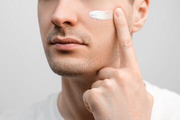 Handsome man with a stubble in a white t-shirt, sample of a face cream on his cheek, smudging with...