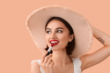 Happy woman in hat adjusting red lipstick on beige background