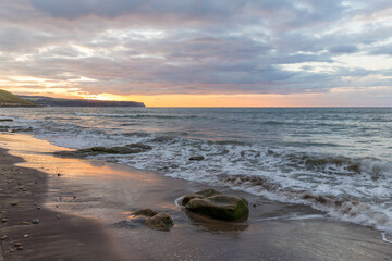 Golden hour sunset at Whitby beach. Warm sky and gentle, smooth waves along a sandy coastline with large rocks.