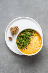 Cream of carrot soup in a white bowl. Carrot soup served in a bowl on a gray background. Bowl of carrot soup with arugula leaves on concrete table