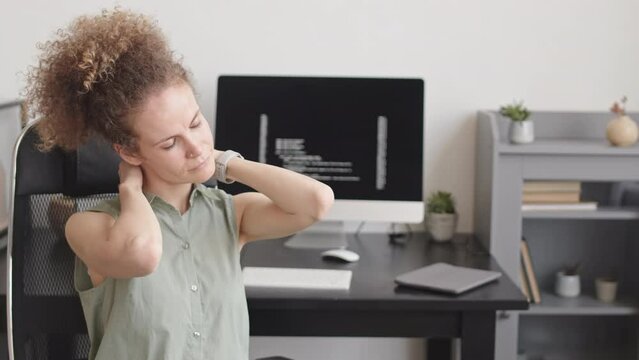 Waist up slowmo of female programmer stretching her neck to relax after desk work sitting on chair at workplace at home office with program codes on black screen of pc computer