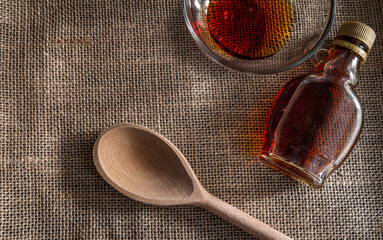Small glass bottle of maple syrup and a wooden spoon on jute material.