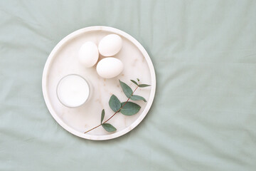 White eggs, marble tray, candle and  branch of eucalyptus on green background. Easter decor. Easter concept. Flat lay, top view. 
