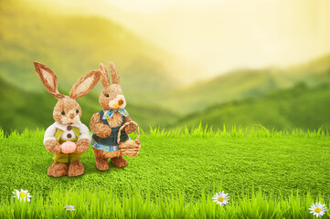 Rabbit dolls made from rice straw stand on green grassland and fresh leafs as frame againt blurred mountain view behind Easter background