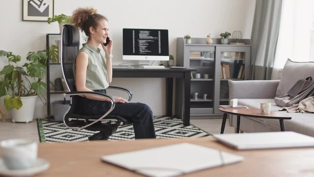 Slowmo of young Caucasian female programmer spinning in chair while having phone conversation working at home office. Program codes appearing on black screen of pc computer in background