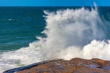 Fototapeta na wymiar Wave breaking over rocks with water splashing with blue sky on a rough sea day in Rio de Janeiro, Brazil