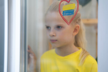 Girl looking out the window, upset and survives the war Ukraine Russia, flag of Ukraine is painted on the window.
