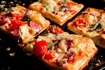 Poster Sliced square pieces of pizza, pie, with tomatoes, sausage, cheese cooked in the oven on a black baking sheet. © Konstiantyn Zapylaie