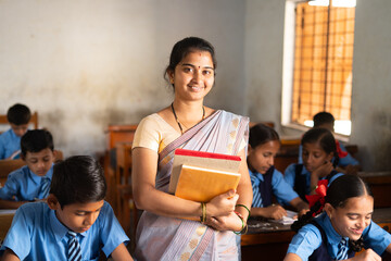 Smiling Teacher with books in hand standing at classroom in between students at classroom - concept of professional occupation, woman empowerment and education - Powered by Adobe