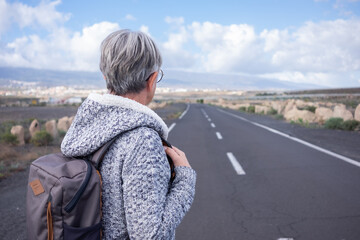 Fototapeta premium Back view of alone senior woman walking outdoor wearing backpack looking at mountain landscape. Elderly mature lady in empty road, mountain and blue sky on background