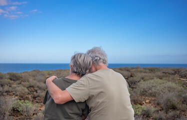 Fototapeta na wymiar Rear view of a tender happy senior couple in love embracing sitting outdoors looking at sea feeling the freedom. Elderly Caucasian man and woman enjoying retirement and healthy lifestyle in nature