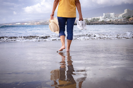 Back View Of Senior Woman Walking Along The Beach With Hat And Shoes In Hands. Mature Lady Bare Feet In The Water All Dressed Up. Scenic Nature And Horizon In The Background