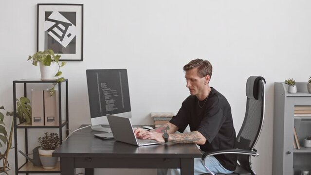PAN medium slowmo of Caucasian male programmer with tattooed arms sitting at desk in modern office typing program code on laptop connected to pc computer