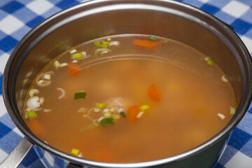 Vegetable soup with meatballs in a stainless steel pan on a table with a checkered tablecloth.