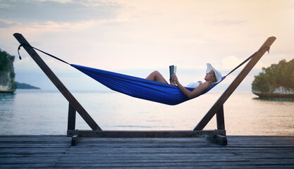 Fototapeta premium Shh, Im on vacation mode. Full length shot of an attractive young woman sitting alone in a hammock and reading a book during a vacation.