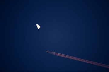 Half moon in the sky with an airplane passing by and leaving its combustion trail, both the trail and the airplane colored by the sunset sunlight