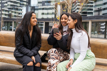 Cheerful Indian women browsing smartphone on bench