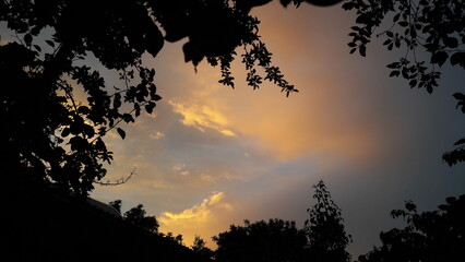 The roof of the house and trees silhouettes against background sunset, evening blue sky with textural clouds