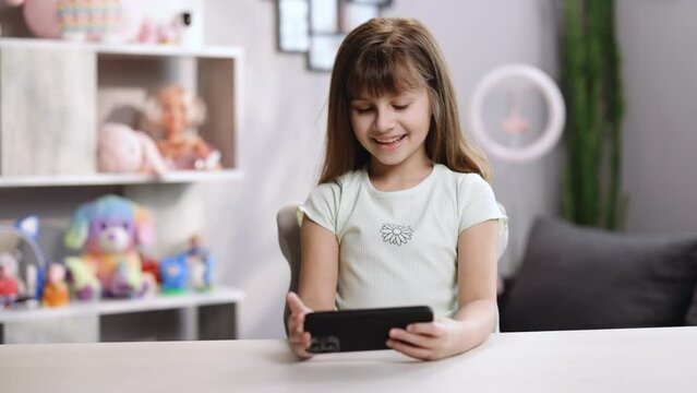 Curious cute preschool kid girl using digital phone technology device sitting in chair alone. Small child hold smartphone surfing internet play game at home. Children tech addiction concept.