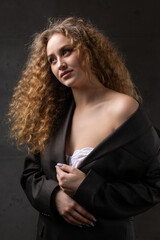 Portrait of a young red-haired curly sexy girl in a jacket with long hair in the studio. Shows off her curvaceous breasts in a bra.