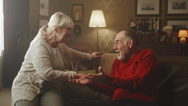 Senior husband in casual clothes telling funny story to laughing wife while relaxing on settee in cozy living room in weekend morning at home