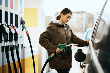 Young woman using fuel nozzles while refuelling gas tank at petrol station.