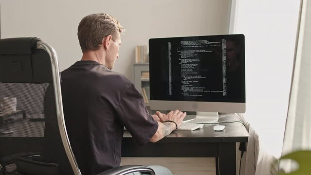 Rear view medium slowmo of young Caucasian software developer in black tee typing program code on pc computer while working from home