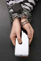 Prisoner holding smartphone in chained hands at black table, top view. Internet addiction