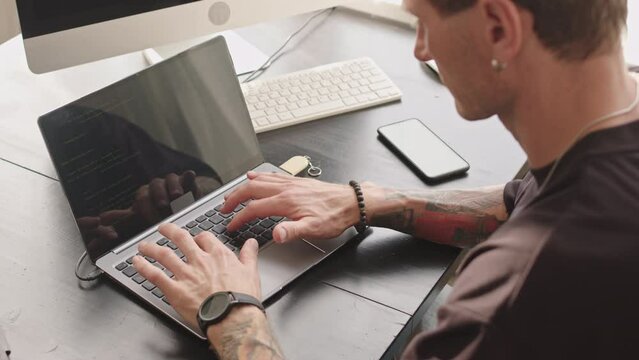 From above medium closeup with slowmo of tattooed Caucasian man working with program code on laptop developing software