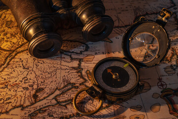 Image of an old compass with a cracked glass and binoculars sitting on map cast in warm candle light.