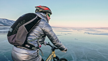 Woman is riding bicycle on the ice. Girl is dressed in a silvery down jacket, cycling backpack and helmet. Ice of the frozen Lake Baikal. Tires on bike are covered with spikes. Traveler is ride cycle.