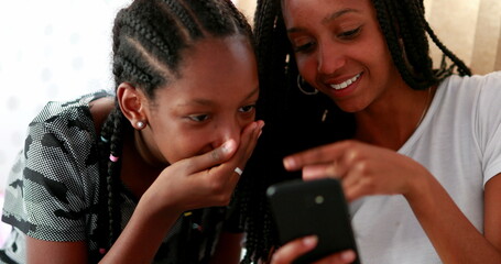 Two teen African girls reacting with shock to news on smartphone. Sisters covering mouth with hands amazed