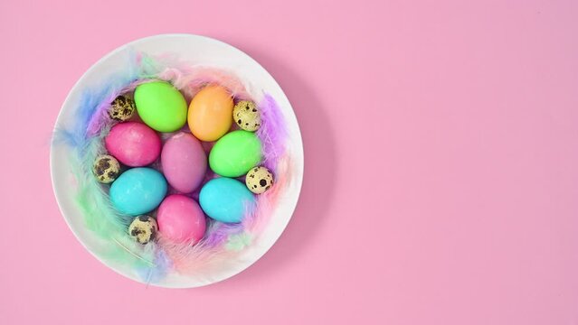 Pastel colorful eggs an quail eggs in white bowl with feathers on pastel pink background. Stop motion