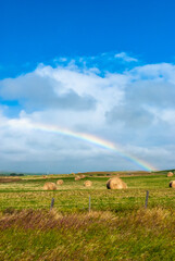 A rainbow arcs across a rural prairie field strewn with round hail bails.