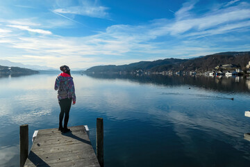 Fototapeta na wymiar Happy woman standing on the pier at beautiful Woerthersee in Poertschach, Carinthia, Austria. Scenic lake landscape surrounded by Karawanks Alps. Fresh and clean air. Reflection in Lake Woerth