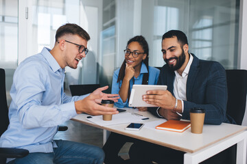 Happy multiracial male and female colleagues watching funny video on modern touch pad technology using 4g wireless during work break in office interior, cheerful employees smiling while browsing web