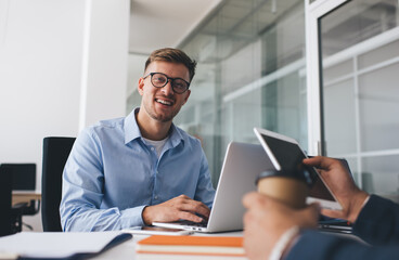 Portrait of cheerful male employee in classic eyewear for vision protection smiling at camera during working process with cropped colleague at table desktop, successful man in optical spectacles