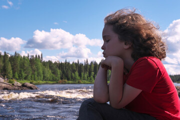 Cute boy sitting on the bank of rapid river on sunny summer day.