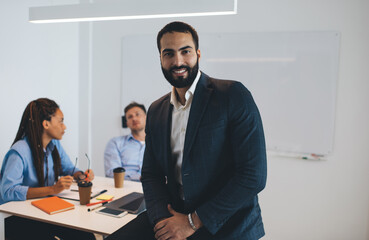 Portrait of happy businessman in formal jacket smiling at camera in office with partners on blurred background, successful Middle Eastern entrepreneur posing during working time in corporate company