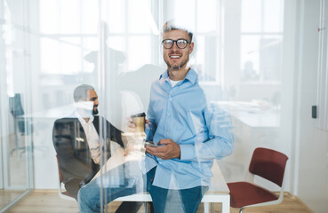 Naklejka na ściany i meble Half length portrait of successful male employee in smart casual clothes smiling at camera during coffee break in office interior, happy businessman in spectacles holding takeaway cup in workspace