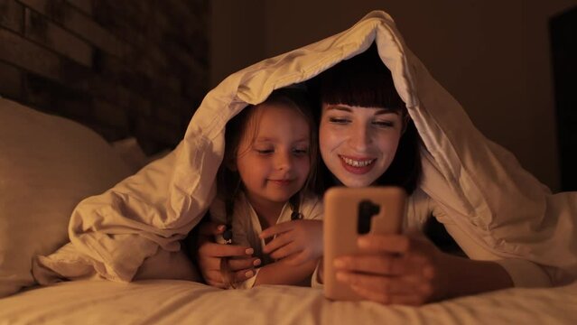 Smiling young woman lying on cozy bed under blanket with her cute daughter, showing mobile application, video game or online cartoons, spending evening time in bedroom.