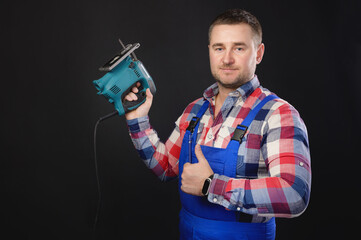 Portrait of a caucasian male repairman in a work uniform with an electric jigsaw in his hand. Studio portrait of an artisan businessman Showing thumbs up