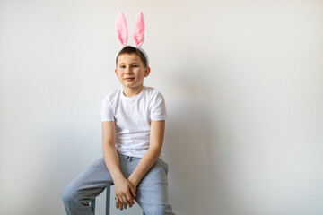 Funny tired boy is wearing bunny ears and sitting on a wooden chair over white background. Preparing for the holiday is a joy for kids. Little child celebrate Easter at home.