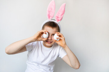 Playful boy with bunny ears keeping white eggs near eyes on white background. Happy child boy is wearing bunny ears and white t shirt.Preparing for the holiday is a joy for kids.